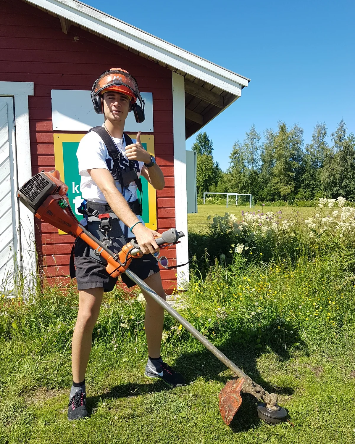 Asensio con casco de protección y desbrozadora, delante de una caseta de madera roja en Finlandia